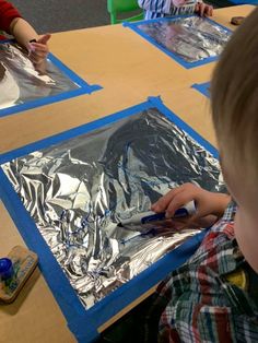 two children sitting at a table with tin foil on them and one child using scissors