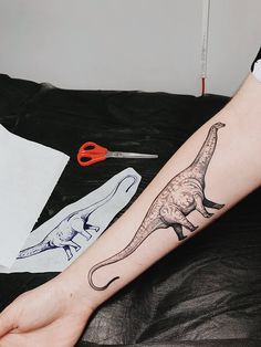 a person with a dinosaur tattoo on their arm next to a pair of scissors and paper