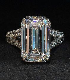 an emerald cut diamond ring with diamonds around it's shoulders and sides, on a black surface