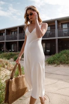 a woman in a white dress is holding a straw bag and talking on the phone