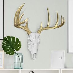 a white and gold deer skull mounted on the wall next to a green leafy plant