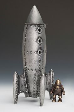 a metal model of a rocket ship with eyeballs on its side and a small figure next to it