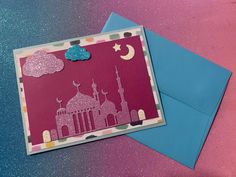 a pink and blue greeting card with a mosque on it, sitting next to an envelope