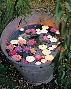 candles floating in a bucket filled with water surrounded by pink and red flowers on the ground