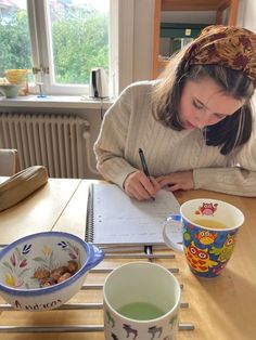 a woman sitting at a table writing on a piece of paper next to two cups