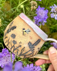 a hand holding a ceramic mug with flowers in the background and purple flowers around it