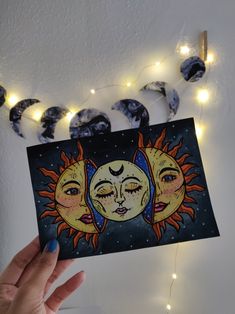 a hand holding up a card with two sun and moon faces on it in front of string lights