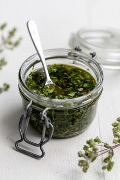 a glass jar filled with pesto next to a metal spoon and sprig of parsley