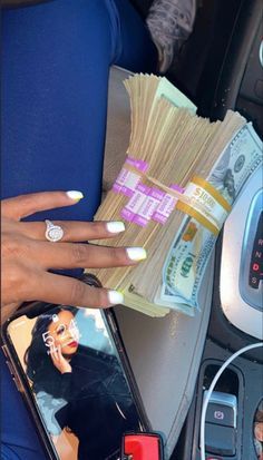 a woman's hand holding money in front of her cell phone and car dashboard