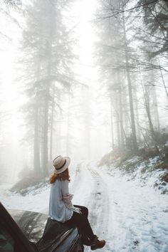 a woman sitting on top of a car in the snow next to some pine trees