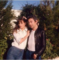 a man and woman standing next to each other in front of some bushes with trees behind them