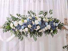 an arrangement of blue and white flowers in front of a curtain with greenery on it