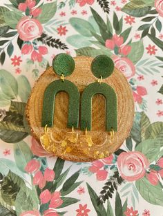 two green earrings sitting on top of a piece of wood in front of floral wallpaper