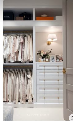 an open closet with white drawers and clothes hanging on the shelves, along with a lamp