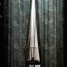 a chair sitting in front of a window next to a flowered curtain with a vase on it