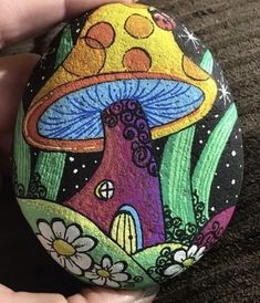 someone is holding a painted rock with a mushroom house in the middle and flowers around it