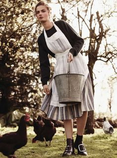 a woman in an apron is holding a bucket and chickens are walking by her on the grass