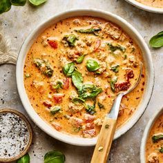 three bowls of soup with basil, tomatoes and other toppings next to a wooden spoon