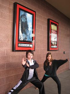 two people standing in front of a brick wall with posters on the wall behind them