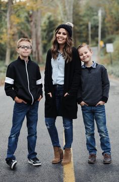 a woman and two boys standing in the middle of an empty road with trees behind them