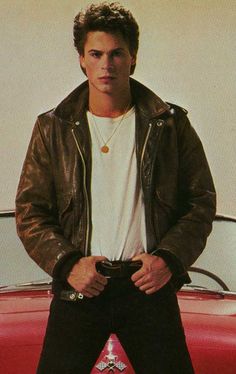 a man standing in front of a red car wearing a leather jacket and white t - shirt