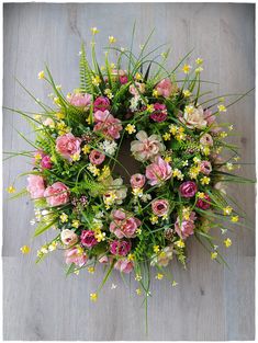 a wreath with pink and white flowers on a wooden floor next to green grass, daisies and yellow flowers