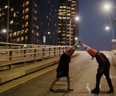 two people with traffic cones on their heads are crossing the street at night in front of tall buildings