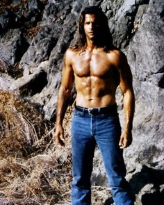 a man with no shirt standing in front of some rocks and grass, posing for the camera