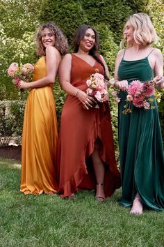 three women in long dresses are posing for the camera with flowers and greenery behind them