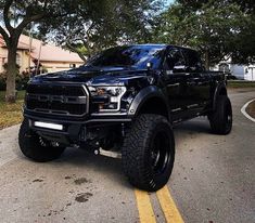 a large black truck parked in a parking lot next to some trees and bushes on the side of the road