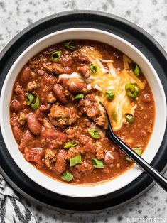 someone is holding a spoon in a bowl of chili and meat soup with cheese on top