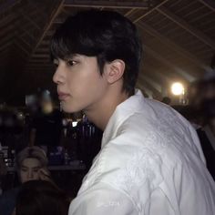 a man with black hair wearing a white shirt and looking off into the distance while standing in front of a group of people