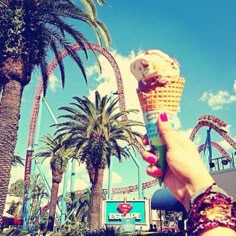 a hand holding an ice cream cone in front of a roller coaster and palm trees