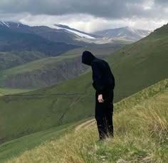 a person in a black hoodie stands on a grassy hill with mountains in the background