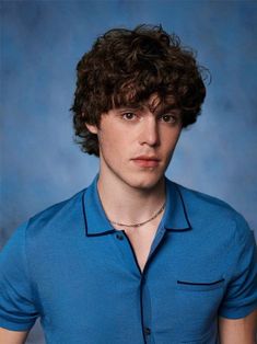 a young man with curly hair wearing a blue shirt and looking at the camera while standing in front of a blue backdrop