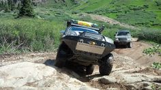 two vehicles driving down a dirt road in the mountains with grass and trees behind them