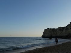 a person walking on the beach near some water and cliffs in the distance with a bird flying overhead