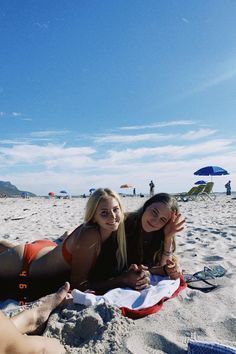 two women laying on the beach with umbrellas in the background and people standing around