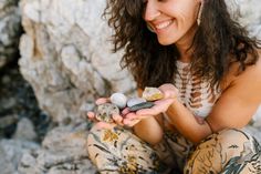 a woman is sitting on the rocks and holding some rocks in her hands, smiling