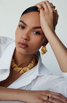 a woman in a white shirt and gold jewelry is posing for the camera with her hand on her head