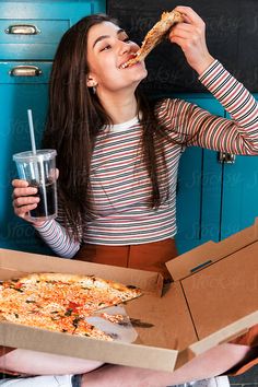 a woman eating pizza while holding a drink in her hand by an open box of pizza