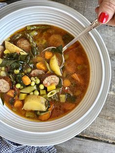 a person holding a spoon over a bowl of soup with meat, vegetables and potatoes