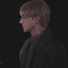 a man with blonde hair wearing a black jacket and looking off into the distance while standing in front of a dark background