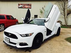 a white mustang parked in front of a building with its doors open and the hood up