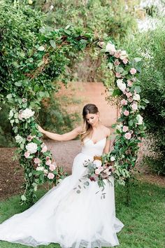 a woman in a wedding dress is sitting under an arch made of flowers and greenery