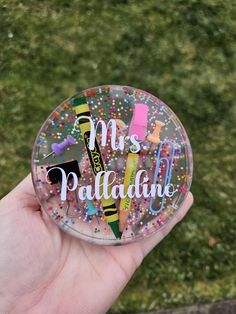 a hand holding a glass magnet with the words miss palladano on it and some crayons