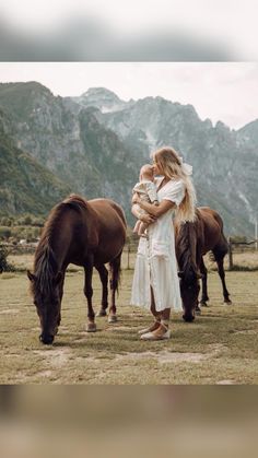 a woman holding a baby standing next to two horses in a field with mountains in the background