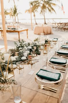 a table set up with place settings and greenery for an outdoor wedding reception on the beach