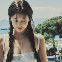 a woman with long black hair standing next to the ocean looking down at her cell phone