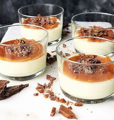 four desserts are sitting in small glass bowls on a white tablecloth with chocolate pieces scattered around them